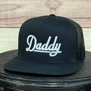 Puede incluir: Gorra de camionero negra con visera curva y parte trasera de malla. La palabra "Daddy" está bordada en escritura blanca en la parte delantera. La gorra está sobre una superficie oscura.