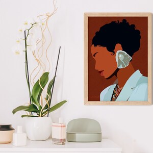 May include: A framed art print featuring a stylized illustration of a person with dark hair and skin, wearing a light blue blazer and a large, shell-shaped earring. The background is a warm brown, and the print is in a light wood frame. A white orchid plant is in the background.