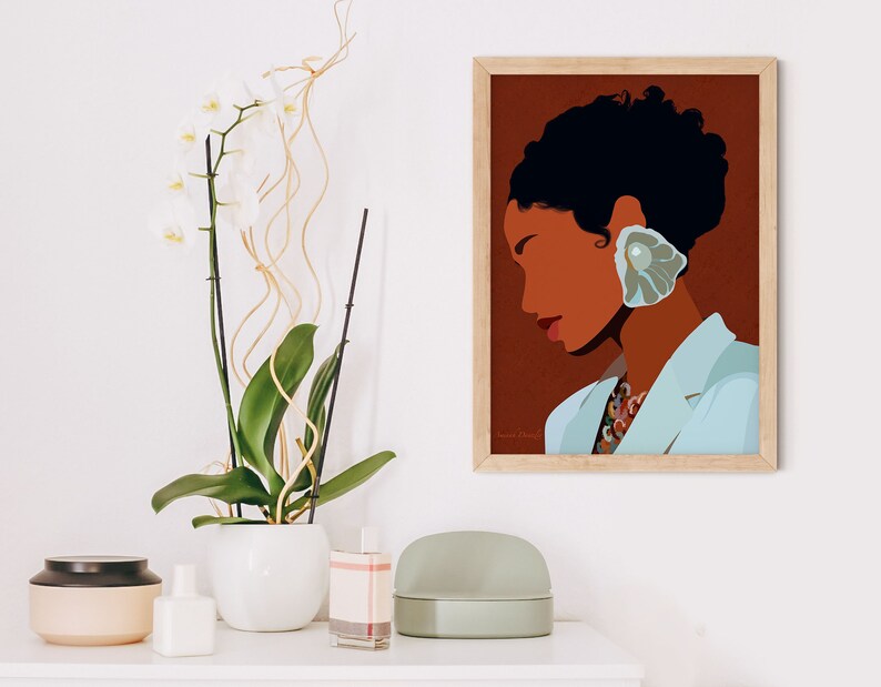 May include: A framed art print featuring a stylized illustration of a person with dark hair and skin, wearing a light blue blazer and a large, shell-shaped earring. The background is a warm brown, and the print is in a light wood frame. A white orchid plant is in the background.