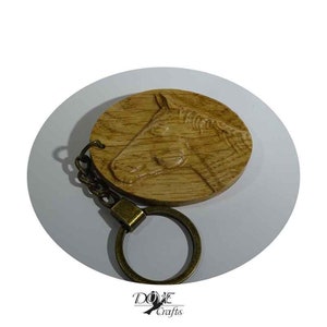 May include: A wooden keychain featuring a detailed relief carving of a horse's head. The keychain has a brass-colored ring and chain. The wood has a natural grain and a warm, light brown color. The keychain is a handcrafted item.