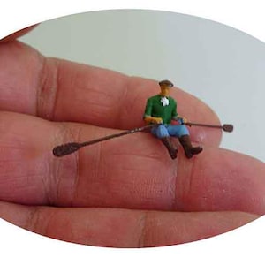 Pode incluir: Uma figura em miniatura de uma pessoa remando em um barco. A pessoa está vestindo uma camisa verde, calças azuis e um chapéu marrom. O barco é feito de madeira e tem dois remos.