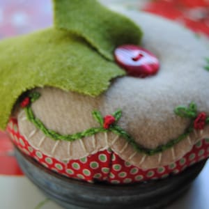 May include: A handmade pincushion with a festive design. It features a beige felt base, a red button, and green felt accents. The base is decorated with a red and green polka dot fabric and a green embroidered garland.
