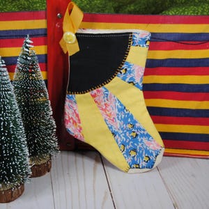 May include: A handmade Christmas stocking with a black top, yellow body, and colorful patterned stripes. A yellow ribbon and button are attached. Three small green Christmas trees with white snow-like accents are in front of a striped red, yellow, and blue container.