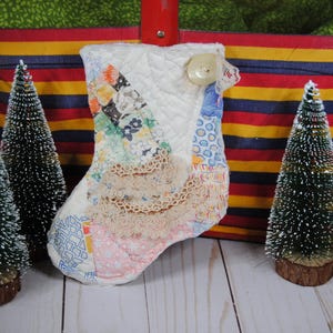 May include: A white quilted Christmas stocking with a lace trim and a variety of fabric scraps sewn onto it. The stocking is decorated with a button and is sitting on a red, yellow, and blue striped surface.