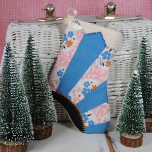 May include: A blue and pink fabric Christmas stocking with a white wicker basket in the background. Two small artificial snow-covered pine trees are on either side of the stocking.