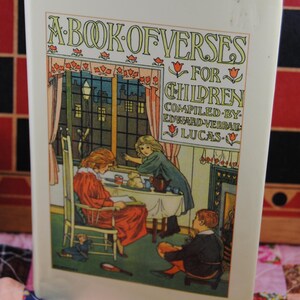 May include: A vintage book titled "A Book of Verses for Children" with a cream-colored cover. The cover features a colorful illustration of children indoors. The book is resting on a patchwork quilt with a wooden ruler in the foreground.