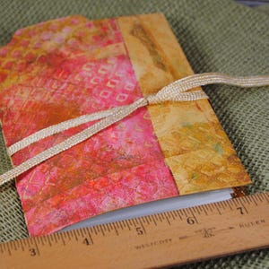 May include: A small, handmade journal with a colorful, abstract cover. The cover features a mix of pink, orange, and yellow hues with a geometric pattern. A beige ribbon is tied around the journal, and a wooden ruler is placed below it.