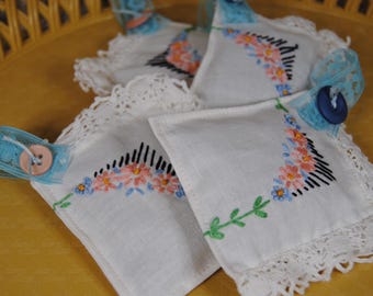 May include: Four white fabric sachets with embroidered floral designs and lace trim. The sachets are filled with dried herbs and have blue buttons attached.