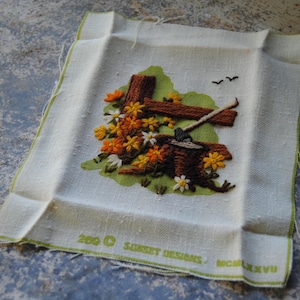 May include: Embroidered fabric square featuring a rustic scene with a brown fence, a tree stump, and colorful flowers in shades of orange, yellow, and white. The fabric has a light green border and the text "Sunset Designs" and "MCMXCVII".
