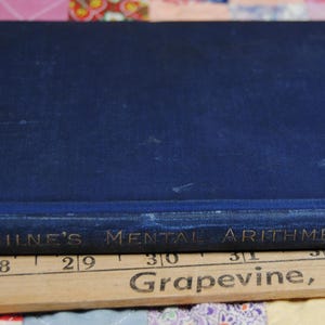 May include: A vintage book titled "Milne's Mental Arithmetic" with a dark blue cover. The book is resting on a wooden ruler with the text "Grapevine, Texas" and measurements in inches. The background is a colorful patchwork quilt.