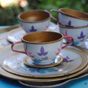 May include: Vintage tea set with white cups, saucers, and a serving tray. The cups and saucers feature a floral design with purple and pink leaves. The cups have red handles and a gold interior. The tray is a light gold color.