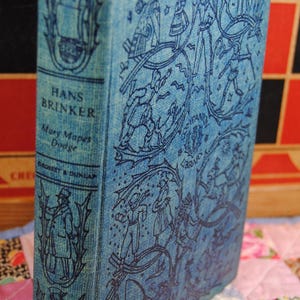 May include: A vintage hardcover book titled "Hans Brinker" by Mary Mapes Dodge. The book has a light blue cover with intricate dark blue illustrations and text. The book is displayed on a colorful patchwork quilt.