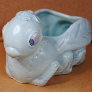 May include: A light blue ceramic planter shaped like a stylized bug with large eyes. The planter has a small opening on the top for planting.