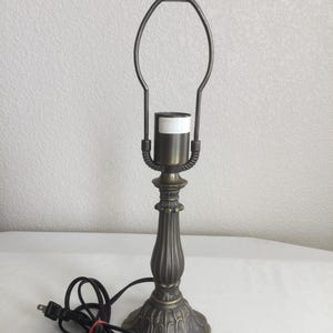 May include: A bronze-toned metal table lamp base featuring a fluted column, a decorative scalloped pedestal, and a harp attachment. The lamp includes a standard light bulb socket and a black electrical cord with a two-prong plug.