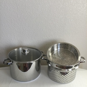 May include: A stainless steel stockpot with a glass lid and a matching stainless steel colander insert. Both pieces feature polished metallic surfaces, side handles, and a cylindrical shape. The colander has a perforated base and sides.