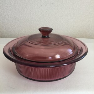 May include: A vintage, round, purple glass casserole dish with a matching lid. The dish has a ribbed exterior and a smooth, rounded rim. The lid features a central knob and a radial pattern. A classic piece for baking and serving.