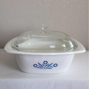 May include: White casserole dish with a clear glass lid. The dish features a blue floral design on the front. The lid has a small, clear handle. A classic piece for serving and baking.