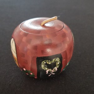 May include: A small, red, wooden apple-shaped box with a painted design of a house with a wreath on the door. The box has a hinged lid and a gold knob.
