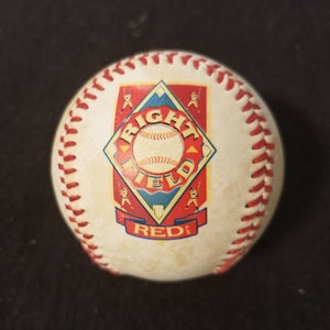 May include: A baseball with red stitching and a printed logo. The logo features a baseball, the words "RIGHT FIELD", and the word "REDS" in a diamond-shaped design with baseball player figures.