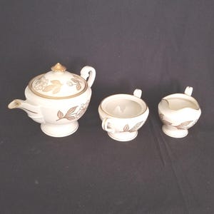 May include: A three-piece porcelain tea set featuring a teapot, a sugar bowl, and a creamer. Each piece is off-white with a metallic gold floral and leaf pattern, gold-trimmed edges, and a gold finial on the teapot lid, all set on a pedestal base.