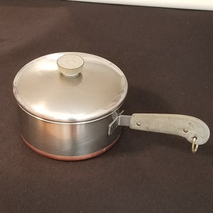 Puede incluir: Una cacerola de acero inoxidable con tapa y asa. La olla tiene un fondo de color cobre y un asa gris. La tapa tiene un pomo gris. El asa tiene un soporte metálico fijado a la olla. La cacerola está sobre una superficie oscura.