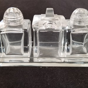 Może przedstawiać: Zestaw solniczki i pieprzniczki ze szkła przezroczystego z dopasowaną tacką. Solniczki mają kwadratową podstawę i okrągły wierzchołek. Taca jest prostokątna i ma zakrzywiony brzeg.