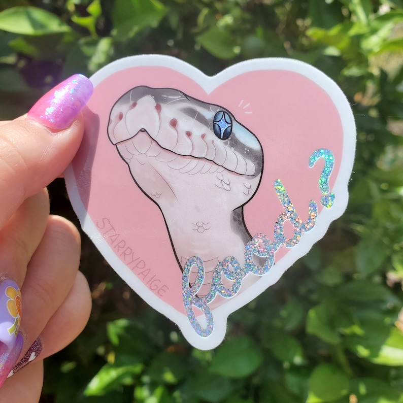 May include: A heart-shaped sticker featuring a stylized snake head with a blue gem-like eye, set against a pink background. The sticker has the text "Good?" in a glittery font and the artist's name, "StarryPaige."