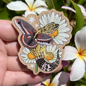 May include: A wooden sticker featuring a colorful gecko with a black and red butterfly perched on its back. The gecko and butterfly are surrounded by white daisies with yellow centers. The sticker has a wood grain background and is perfect for adding a touch of whimsy to any surface.