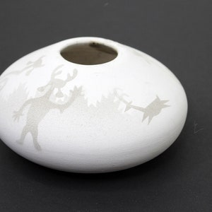 Może przedstawiać: Biały ceramiczny wazon o zaokrąglonym kształcie z małym otworem u góry. Wazon jest ozdobiony wytrawionymi figurami stylizowanych ludzi i drzew. Wazon stoi na czarnej powierzchni.