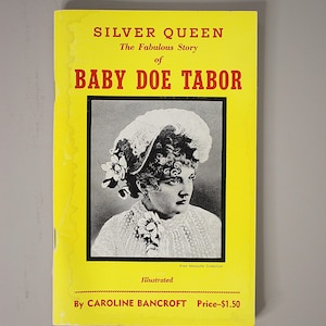 May include: A yellow paperback book titled Silver Queen The Fabulous Story of Baby Doe Tabor by Caroline Bancroft. The cover features a black and white portrait of a person in a decorative hat and lace collar, with red and black text.