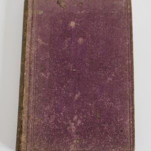 Puede incluir: Un libro antiguo con una cubierta de color morado oscuro y bordes dorados. La cubierta está desgastada y desteñida, mostrando signos de la edad.