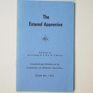 May include: A light blue booklet titled The Entered Apprentice with black text and a Masonic square and compass symbol. The cover states it is authorized by the Grand Lodge A. F. & A. M. of Kansas, compiled by the Committee on Masonic Education, BOOK NO. TWO.