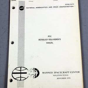 Puede incluir: Un manual vintage del Manned Spacecraft Center, Houston, Texas, con el logo de la NASA. La cubierta blanca presenta el texto "METROLOGY REQUIREMENTS MANUAL" y "NOVEMBER 1970". El documento tiene tres perforaciones en el borde izquierdo.