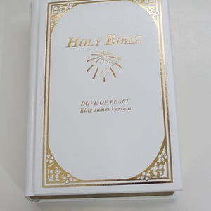 Puede incluir: Biblia de cuero blanco con letras doradas y un diseño de paloma de la paz en relieve dorado. El título dice "Holy Bible Dove of Peace King James Version".