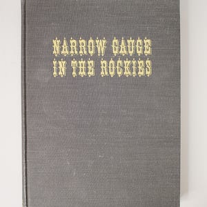 Puede incluir: Un libro de tapa dura con una cubierta de tela gris texturizada. El título NARROW GAUGE IN THE ROCKIES está impreso en letras doradas en la portada. El libro tiene una forma rectangular con un lomo vertical en el lado izquierdo de la cubierta.
