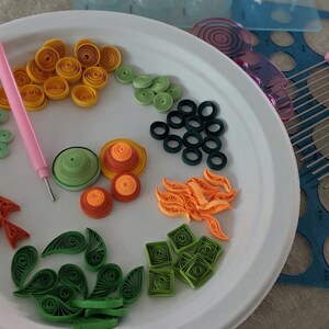 May include: A white plate with various paper quilling designs in different colours, including green, orange, yellow, and red. The designs are arranged in a circular pattern on the plate. There is a pink tool for quilling on the plate.