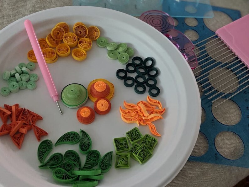May include: A white plate with various paper quilling designs in different colours, including green, orange, yellow, and red. The designs are arranged in a circular pattern on the plate. There is a pink tool for quilling on the plate.