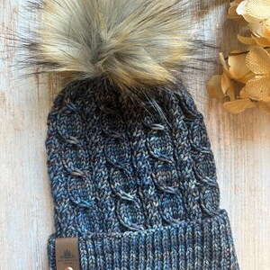 May include: A blue and brown knit beanie with a large faux fur pom-pom. The hat features a cable knit design and a leather tag with a logo. The beanie is approximately 9 inches tall.