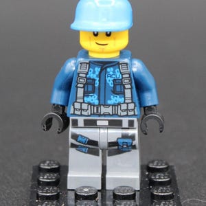 May include: A Lego minifigure with a light blue helmet, dark blue jacket with "ACU" text, gray pants, and black hands. The figure stands on a black base with black studs. The jacket has a camouflage pattern.