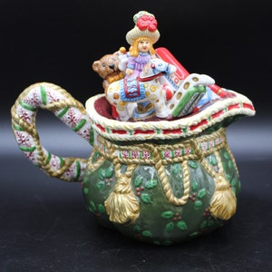 May include: A green ceramic pitcher shaped like a Santa sack with a rope trim and holly accents. The pitcher has a handle and is topped with a girl doll riding a rocking horse, a teddy bear, and other toys.