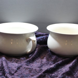 May include: Two white ceramic chamber pots with handles. The pots are sitting on a purple velvet fabric.