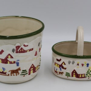May include: Two ceramic containers with a winter village scene. The larger container is a cylinder with a green rim. The smaller container is oval with a handle and a green rim.