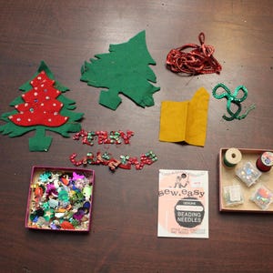 May include: A craft kit with felt Christmas tree shapes, red and green sequins, and a package of beading needles. The kit also includes a small box of assorted sequins and two spools of thread.