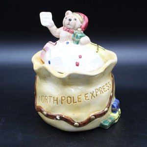 May include: A ceramic Christmas cookie jar shaped like a Santa sack with a teddy bear wearing a Santa hat on top. The sack is decorated with a brown strap and gold studs. The text "NORTH POLE EXPRESS" is printed on the front of the sack.