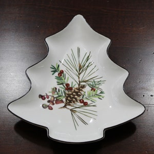 May include: White ceramic Christmas tree-shaped serving dish with a black rim. The dish features a painted design of a pine branch with pine cones and red berries.