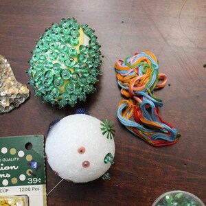 May include: A collection of craft supplies including a green and yellow beaded egg-shaped ornament, a white and pink decorative ball, and a bundle of colorful embroidery floss. Also, a package of "Fashion Trims" with the text "WALBEAD".