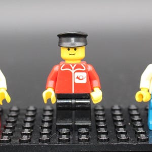 May include: Three Lego minifigures on a black base. The figures are wearing red, black, and blue outfits with matching hats. The figures have yellow heads and smiling faces. One figure has a wrench logo on their outfit.