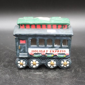 May include: A ceramic train car painted in black, green, red, and white. The train car has a green roof, red and white stripes, and a sign that reads "HOLIDAY EXPRESS".