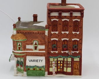 May include: A miniature brick building with a green awning that reads "VARIETY". The building has a red roof and white trim. There are wreaths in the windows and a candy cane in front of the building.