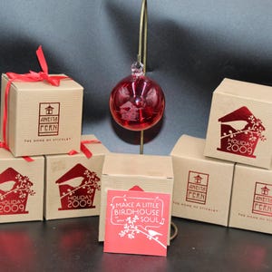 May include: A collection of small, square gift boxes with red ribbon, featuring a red birdhouse design and the text "HOLIDAY 2009." A red glass ornament hangs above the boxes. The boxes are made of brown cardboard.